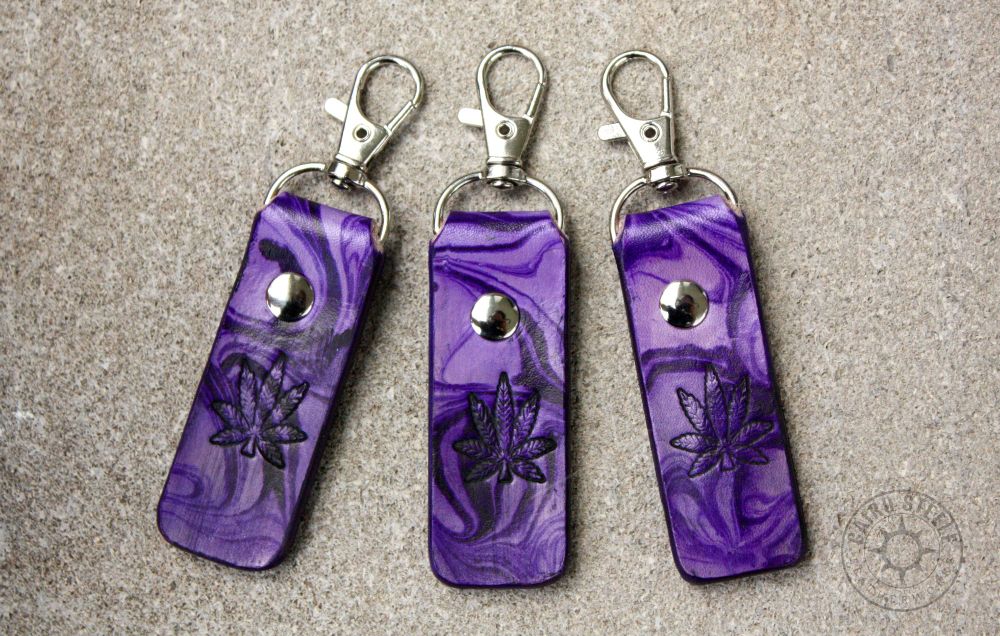 3 leather keychains in purple marble, with a hemp leaf stamped on the front. Each has a silver colored clip and matching rivet.