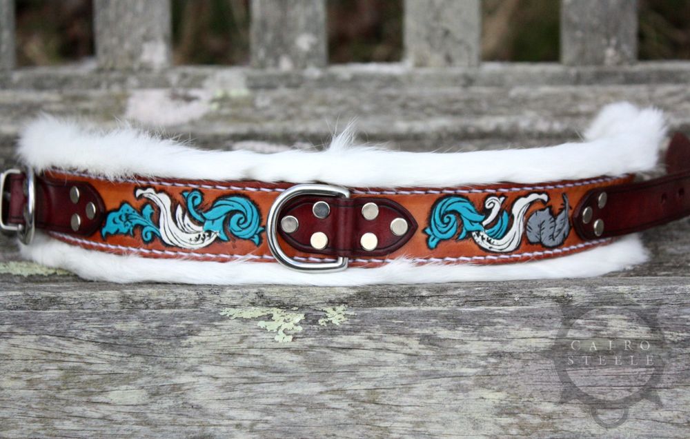 A handmade leather collar. It has a medium brown base strap with tooled scrollwork across the entire body and a feather on one side. The scrollwork is painted white and turquoise, and the feather is metallic silver. The top straps of the collar are dark brown with silver colored hardware. It's lined with super soft white rabbit fur and stitched together with white thread.