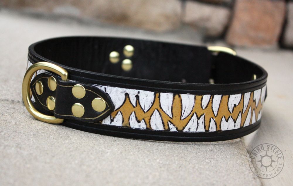 A handmade leather collar. The straps are black, and the base strap has been tooled with snaggley teeth. The teeth are painted white and the area between them is metallic gold. It has solid brass hardware.