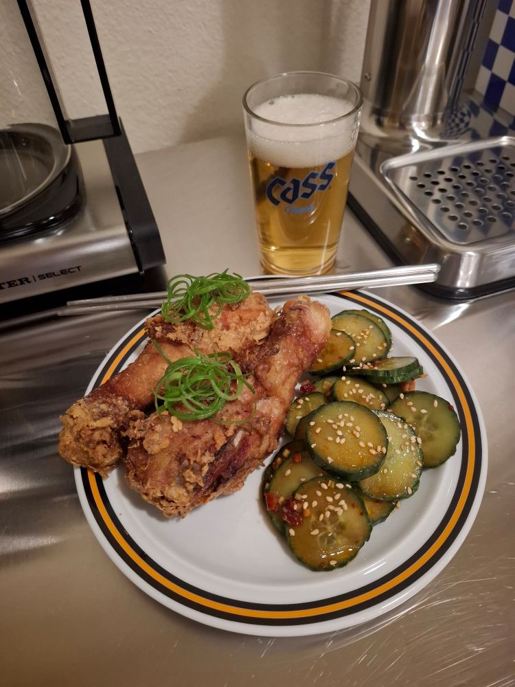 auf kleinem weißen teller zwei hühnerschenkel ayam goreng stil frittierr, daneben ein koreanischer gurkensalat 
das huhn ist mit gelockten frühlingszwiebeln und der salat mit weißem sesam garniert 
dahinter ein kleines bier in einem glas