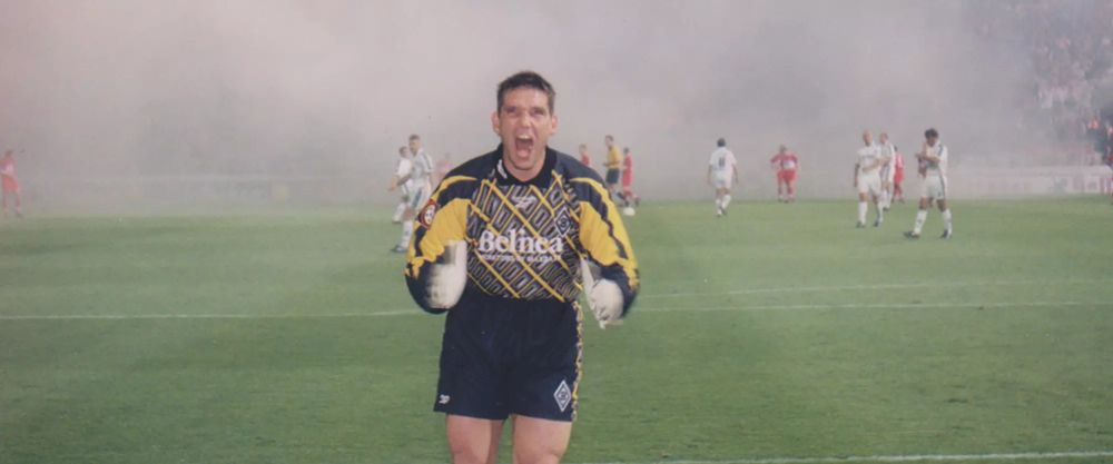 uwe kamps rennt im torwart dress auf die kamera zu und scheint schreiend in seine hände zu klatschen hinter ihm stehen team und gegenspieler im rauch der von der tribüne zieht