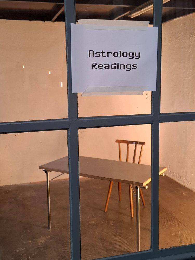 hinter fensterglas ein tisch und ein stuhl in sonst leeren raum mit betonboden 
auf blatt in pixelschrift an der scheibe steht "Astrology Readings"