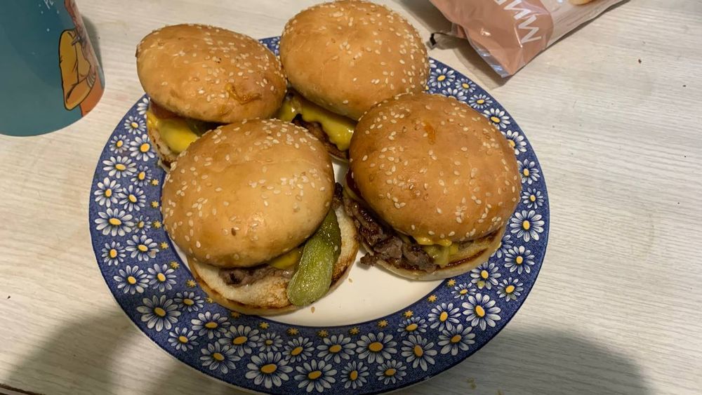 a plate with 4 juicy, thicc burgers residing on it. a pickle is sticking out of one of the burgers