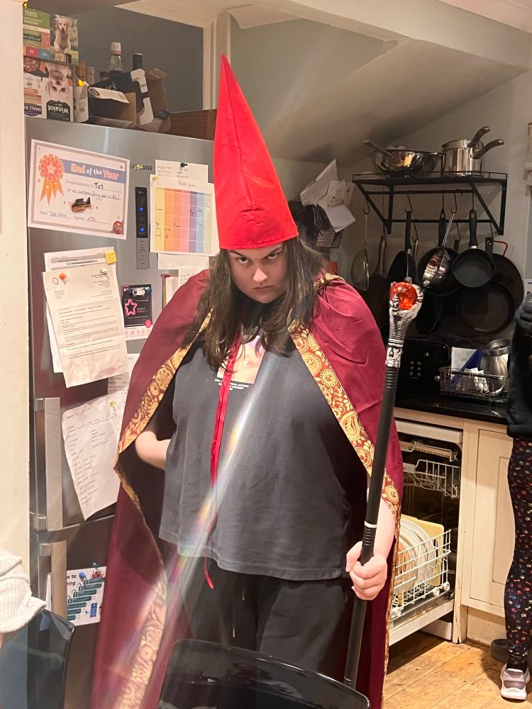 Child1 in robe, Gnome hat and enchanted plastic stick, standing in the kitchen.