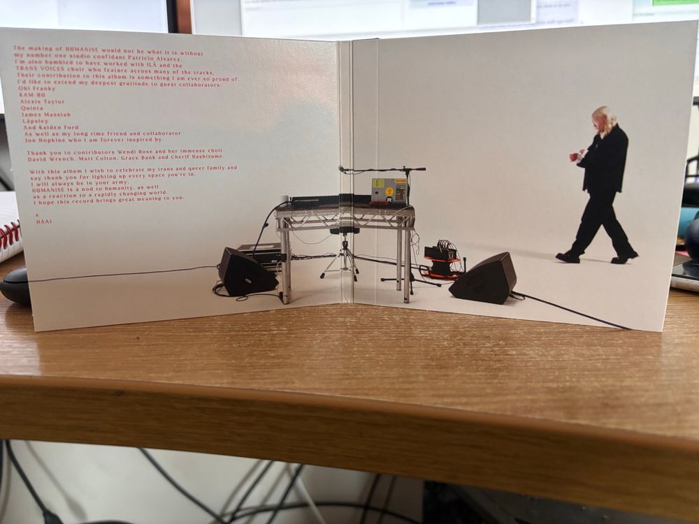 The inside faces of Haai's album Humanise. Featuring the artist wearing a baggy suit against a white background approaching a stand with synth, laptop, mic, stool, power cables and foldback/monitor speakers. 