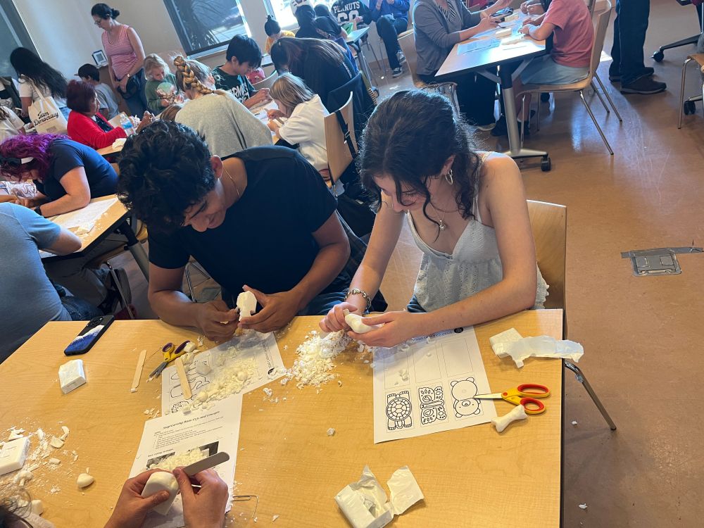A close up of two soap carvers at a table with other soap carvers in the background. 