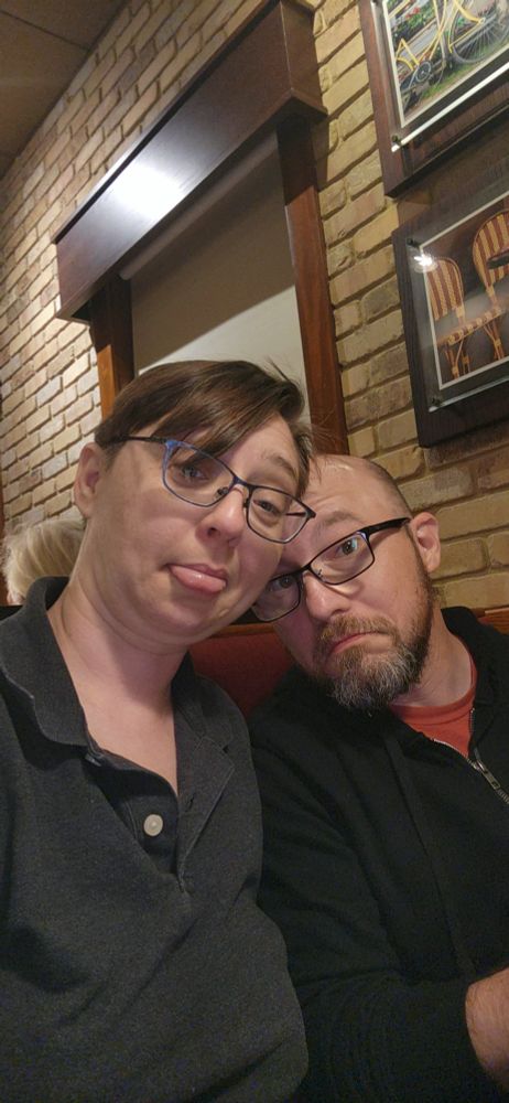 Woman with short hair sitting next to a man with a beard, both making silly faces at the camera