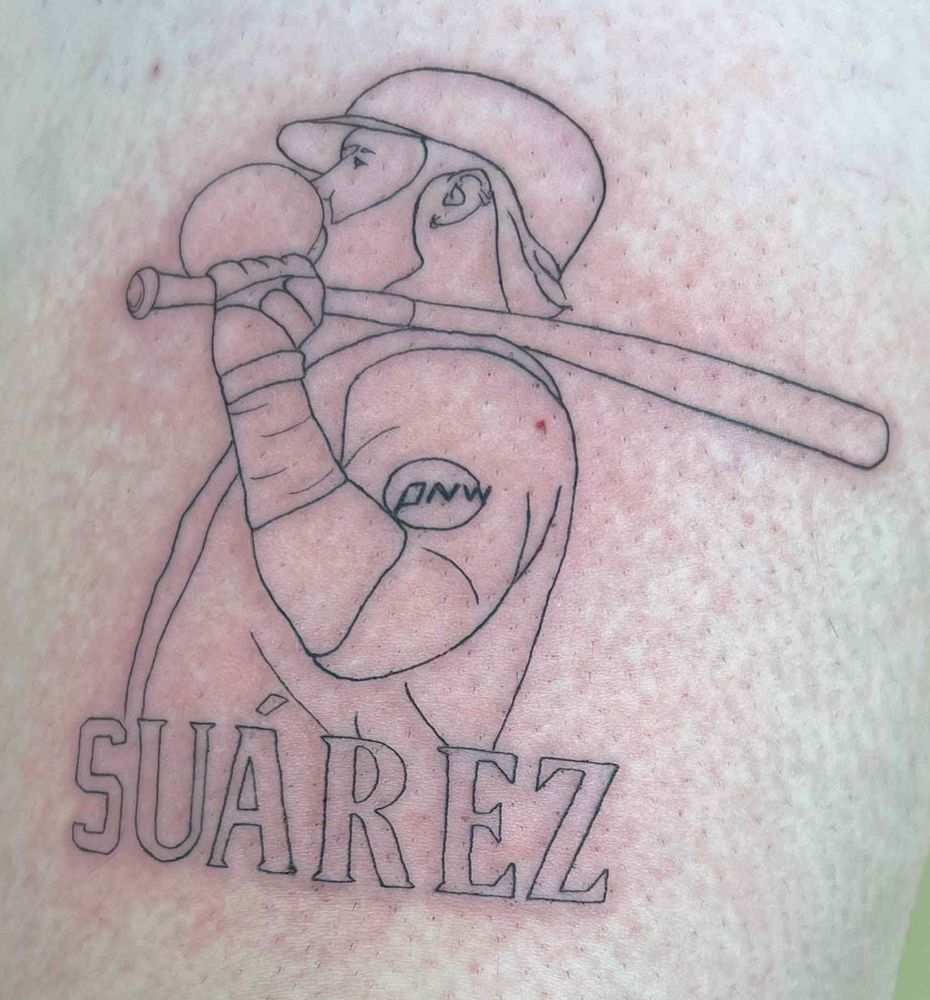 A tattoo of Eugenio Suárez blowing a bubble with his bat over his shoulder. He has on a batting helmet and batting gloves. Below it says Suárez in capital letters. 