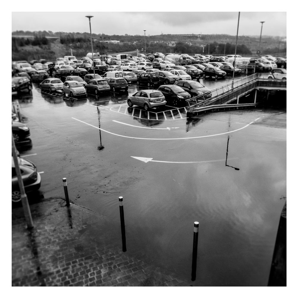 From an elevated position, looking down into a partially flooded car park. The standing water acts like a mirror and reflects an overcast sky. In the foreground there are metal posts whilst in the background there is an expanse of parked cars and some lampposts. 