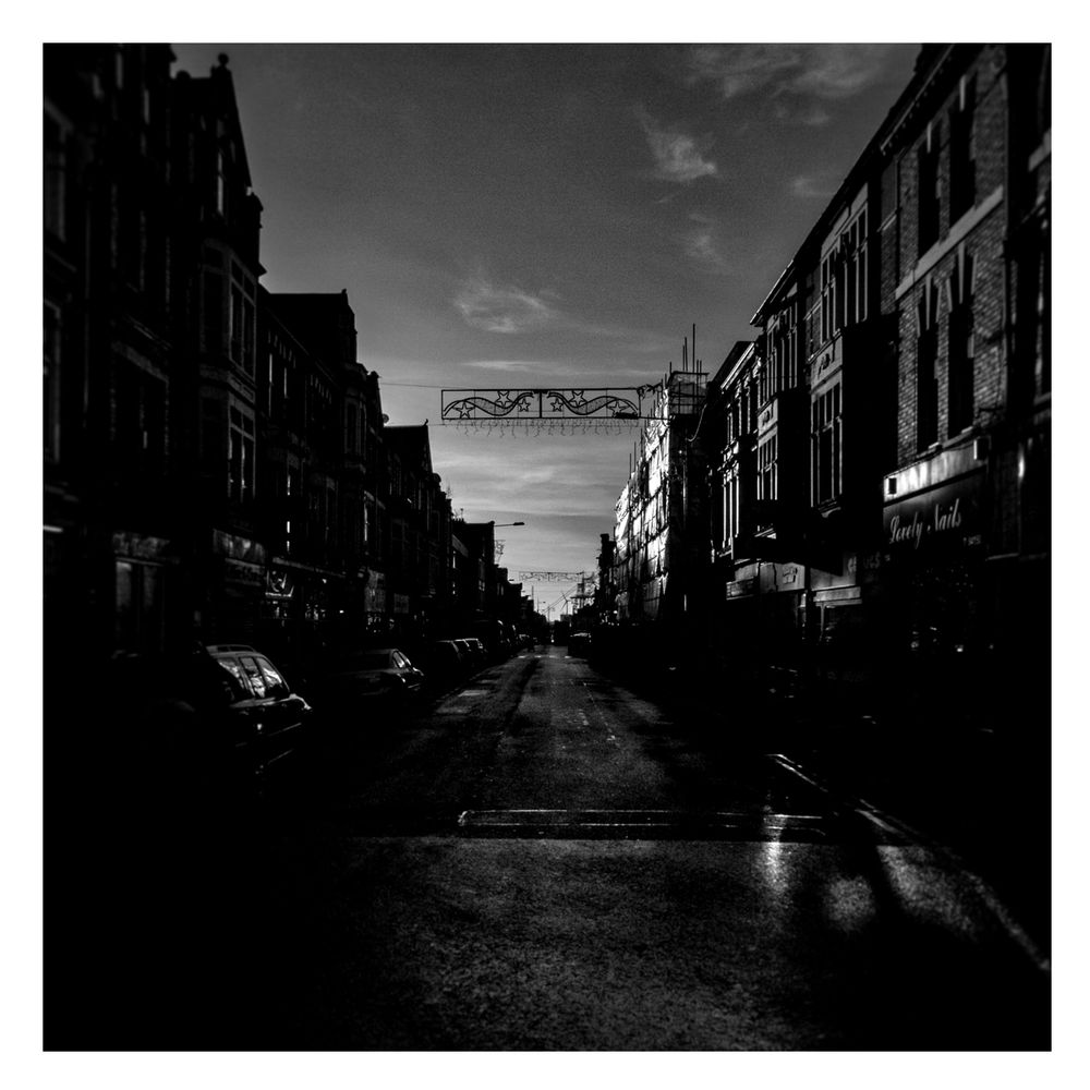A view down a street in deep winter shadow. Hanging between two buildings hangs a tired looking set of Christmas lights. Sunlight picks out the odd window and shop front.