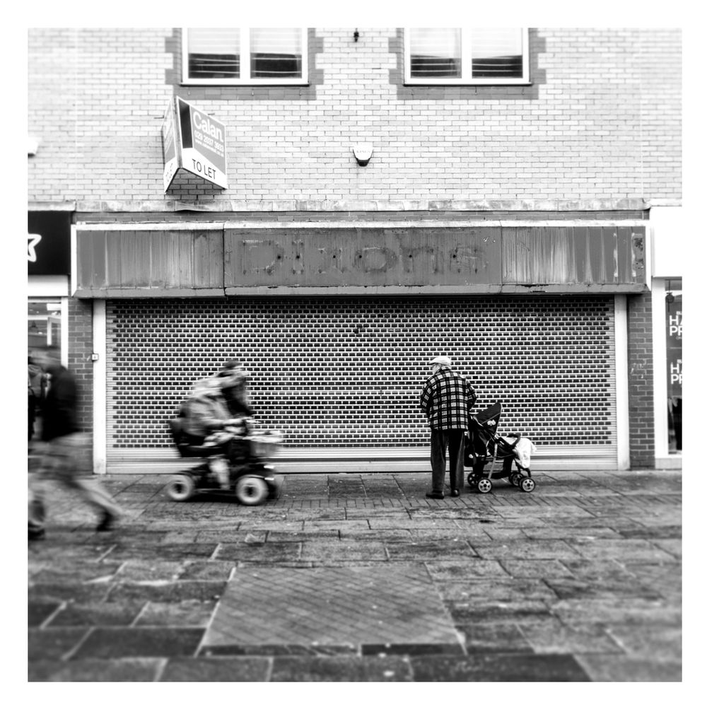 Framed by the outline of a vacant and shuttered shop, there stands a man with a push chair in the right hand side of the frame, looking left towards two people approaching him. One is on a mobility scooter whilst the other walks. They're slightly indistinct due to a low shutter speed which gives the impression that they are moving at speed. The man who they are approaching is stock still, frozen in time.