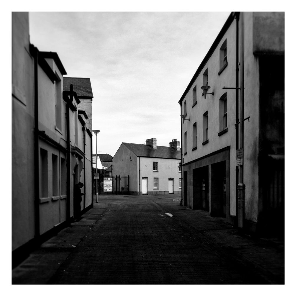 A quiet side street, more flat dreary light and just visible in a door way on the left someone is smoking a cigarette whilst blowing on their phone - maybe an employee from the nearby pub having a break from all the day drinkers. 