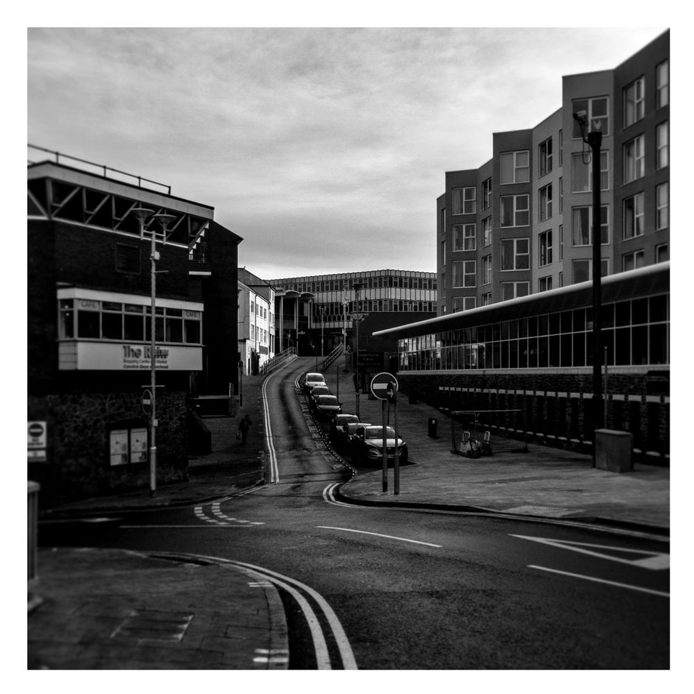 A dreary view towards what was once the Co-op and subsequently what was once Wilko. A road snakes up the middle of the frame towards said empty shop. A row of cars is picked out by the fact and miserable light. On the right of the frame stands a typically bland block of flats, whilst on the left there is the indoor market, which itself is closed now. 