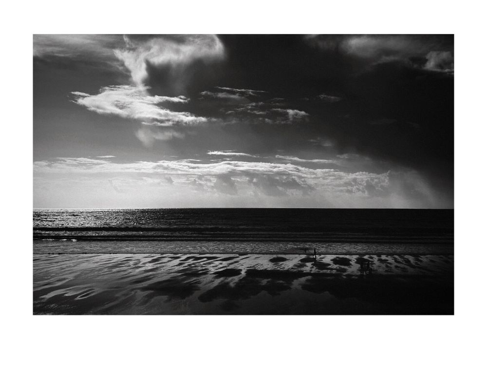 A across the beach at Aberavon, Port Talbot. Looking from left to right the sky starts clear and as we look across the cloud builds up significantly, darkening as we look right. The sand is firm and it looks like the tide has recently gone out - streaks of water run towards the tide. In the distance, whiter clouds can be seen, a promise of a brighter day lurks within. A solitary figure can be seen walking on the beach, no doubt enjoying the serenity of nature's bounty.