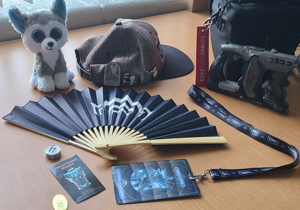 A sprawl of warframe goodies on a table. A hat with an Excalibur pin, a fan with the lotus symbol, a lanyard with a Tennocon general admission, a tag for Tennocon 2025, a slip depicting a Harrow Chassis blueprint, a small piece of warframe platinum, a replica Viper weapon, and a flattened coin depicting a place called The Factory. Alongside all of this is a plush husky used to depict the poster.