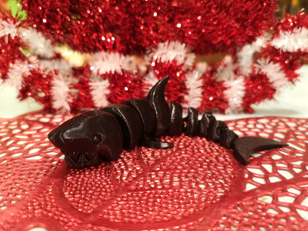 In the Background there is a red and white Christmas decoration used as a prop to create the background. On the table a red Christmas placemat is also used as decor. An all black articulated shark is placed in the center. It is segmented and laying on its stomach.