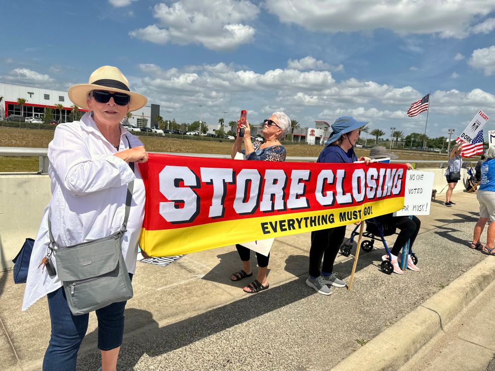 Protestors holding a βStore Closing, Everything Must Goβ banner in front of the Tesla showroom.