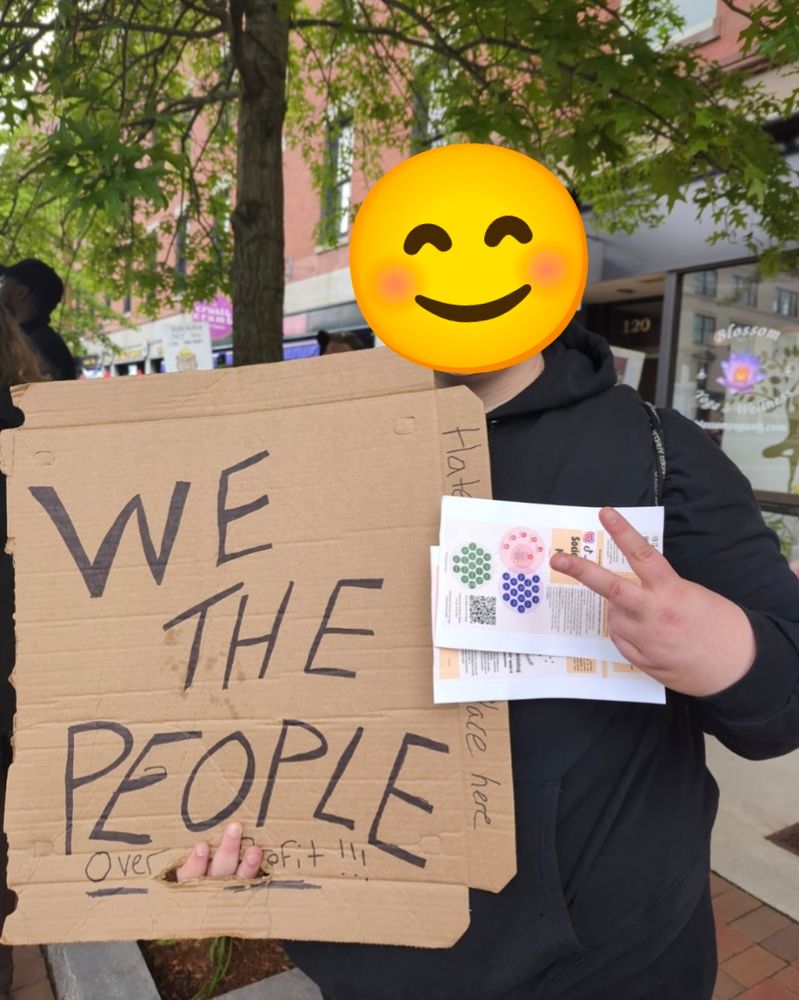 Protester holding a sign that reads We The People Over Profit along with GSUS flyers and a peace sign. A smiling emoji covers the protesters face