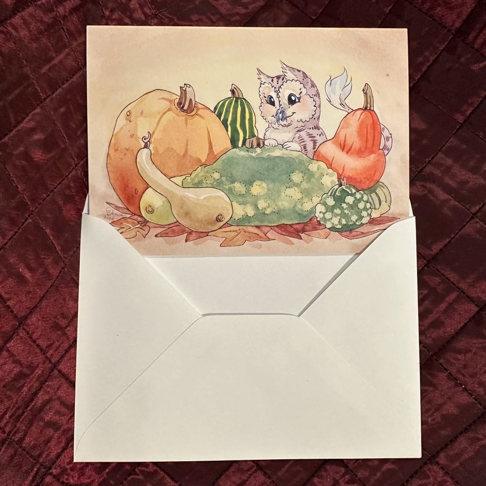 Picture of a greeting card on a dark red background. The card features a cute owl-cat monster with a pile of pumpkins.