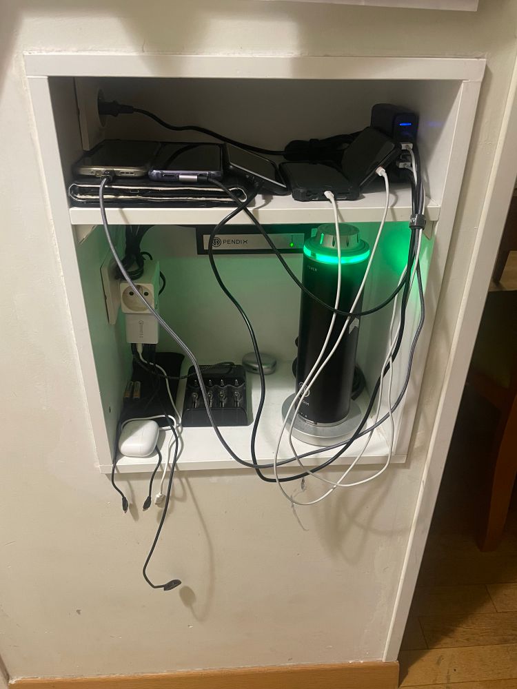 A small alcove with two shelves holding a variety of electronic devices and chargers. Bottom shelf has a large Pendix bike battery. 