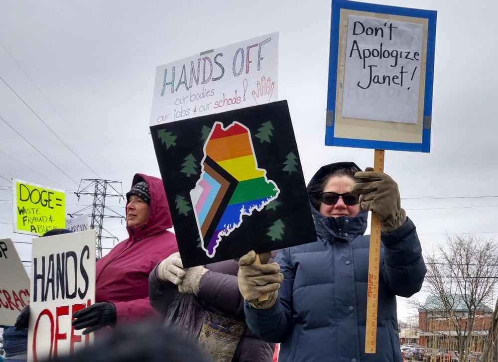People holding up signs at a rally. Some of the signs read: DOGE = Waste Fraud & Abuse; HANDS OFF!; HANDS OFF Our Bodies, Our Jobs, Our Schools; Don't Apologize Janet! and a sign with an image of the state of Maine with a inclusive Pride flag.