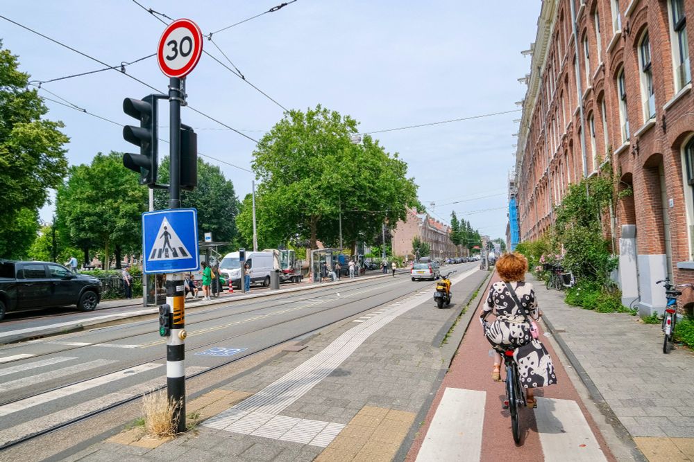 People of all ages and abilities walk, cycle, and drive along the low-traffic streets of Amsterdam, which are lined with buildings, trees, bike infrastructure, and prominent 30 km/hr signage.