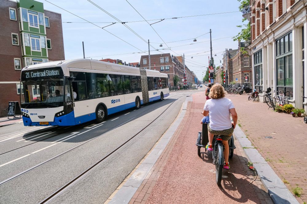 People of all ages and abilities walk, cycle, and drive along the low-traffic streets of Amsterdam, which are lined with buildings, trees, bike infrastructure, and prominent 30 km/hr signage.