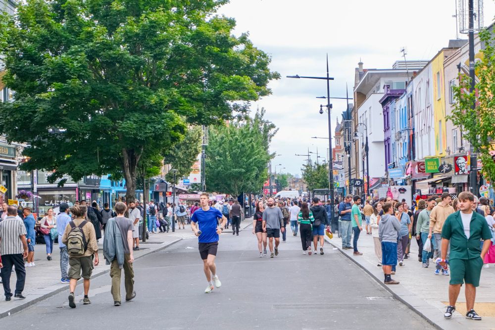 People walk, cycle, sit, and talk along a temporarily car-free stretch of Camden High Street in North London, which is lined with retail shopfronts, trees and flowers, terraces, bike parking, and benches.