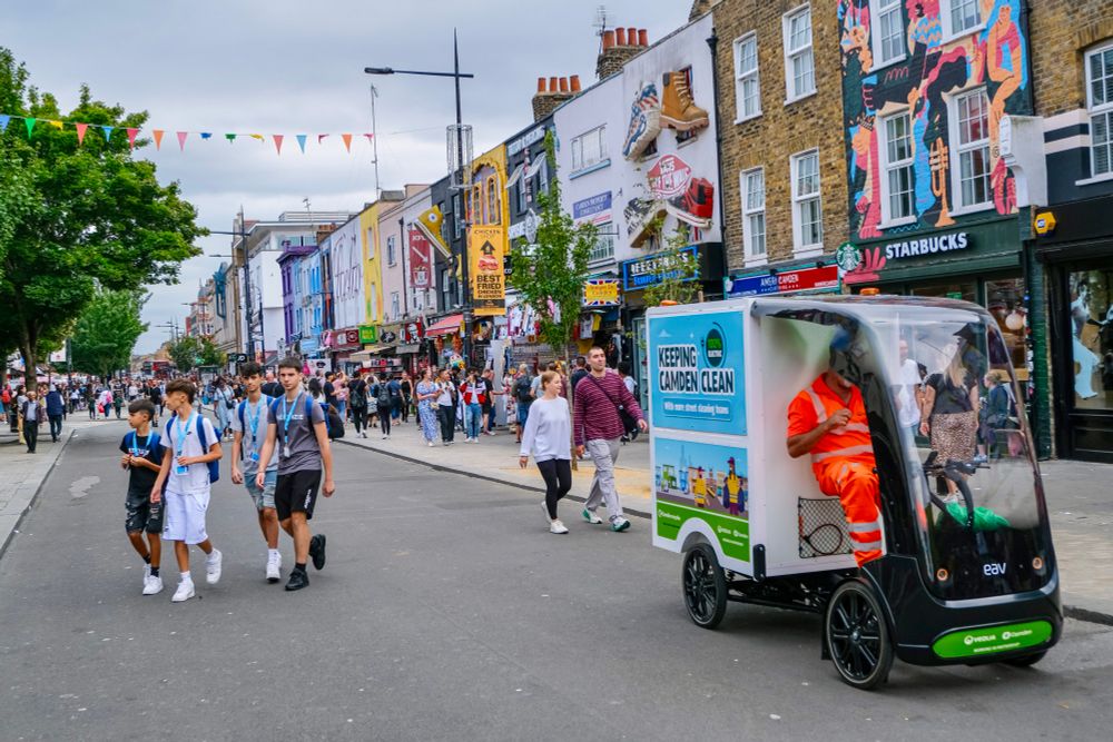People walk, cycle, sit, and talk along a temporarily car-free stretch of Camden High Street in North London, which is lined with retail shopfronts, trees and flowers, terraces, bike parking, and benches.