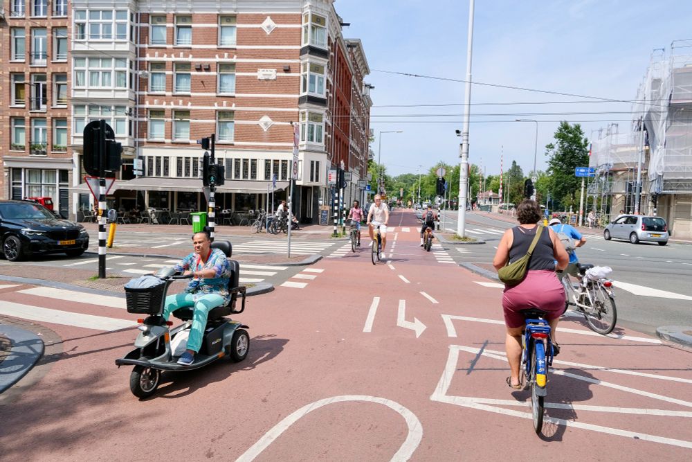People of all ages and abilities walk, cycle, and drive along the low-traffic streets of Amsterdam, which are lined with buildings, trees, bike infrastructure, and prominent 30 km/hr signage.