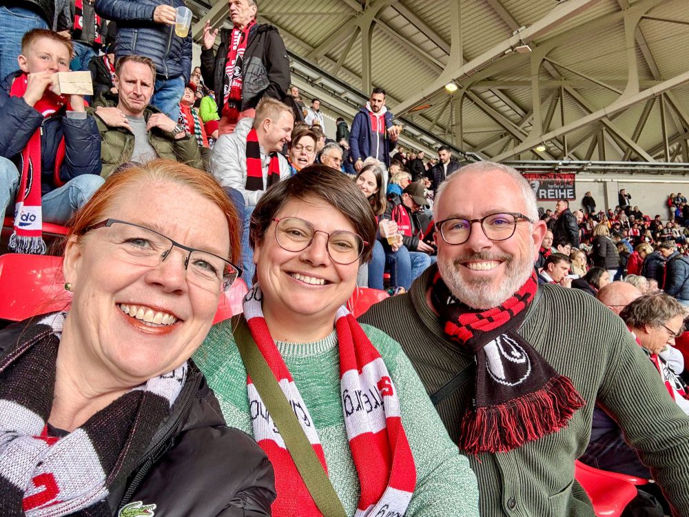 Three middle aged adults wearing red and white scarves pose for a selfie during a SC Freiburg football match.
