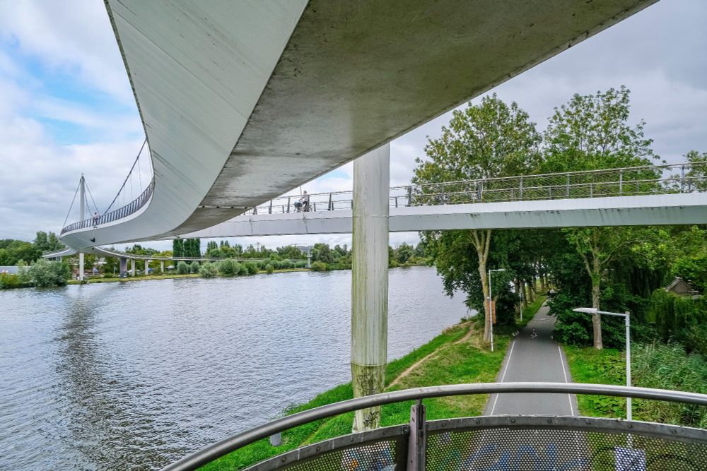 Cyclists and pedestrians in casual autumn attire travel across the Nesciobrug in Amsterdam, a white steel cable-suspension bridge for non-motorised traffic stretching high across the Amsterdam-Rijn Canal.