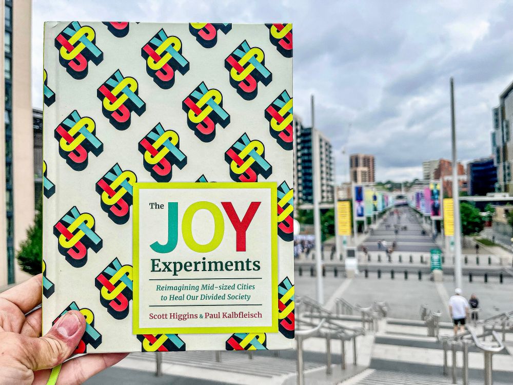 A hand holds a book, “The Joy Experiments”, in front of a long pedestrian street in London.