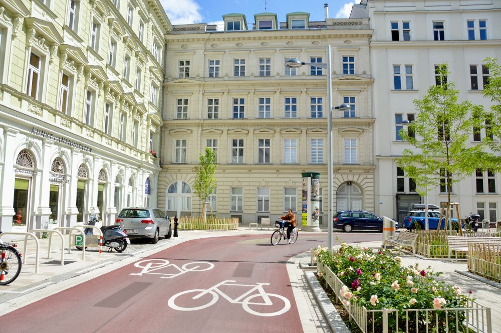 People in casual spring attire cycle on Argentinierstraße in Vienna—a red asphalt “bicycle street” lined with four-storey buildings, trees and plants, and wooden benches. 