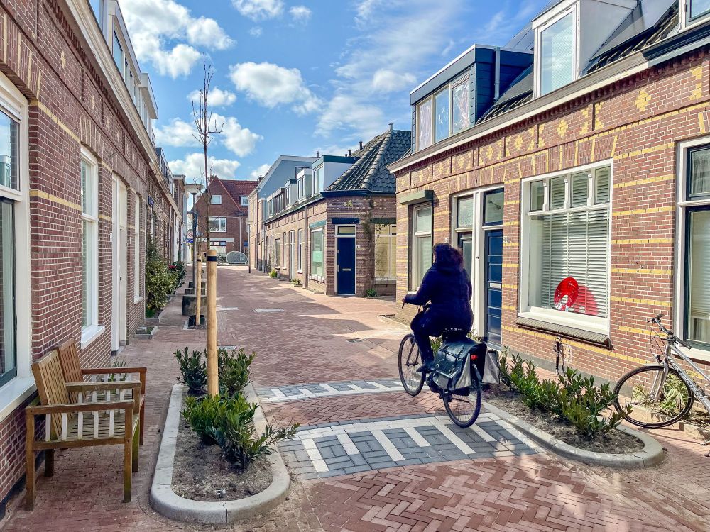 People walk along a “woonerf” or “living street” lined with trees, plants, lampposts, benches, and two-storey brick buildings in Delft, The Netherlands.