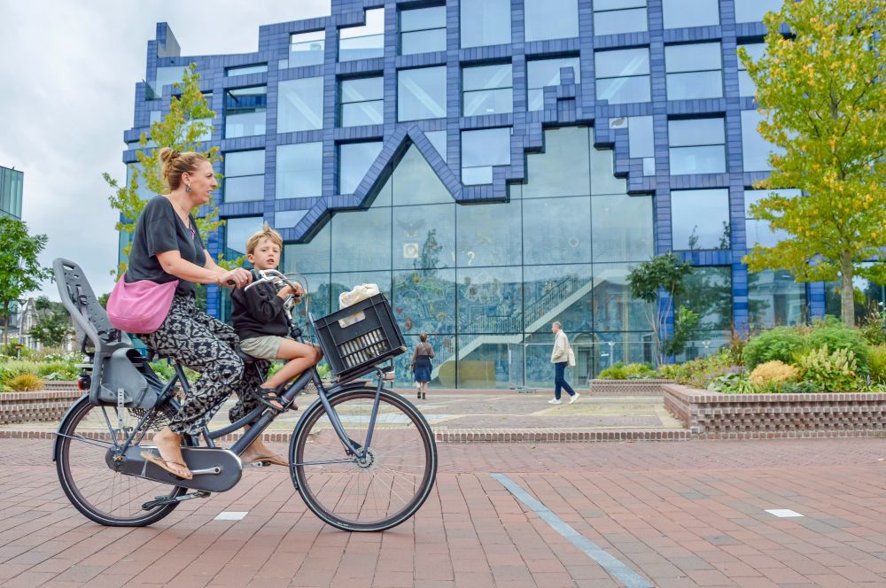 People of all ages and abilities ride bicycles along the red asphalt cycling paths and cycling streets of Delft, which are lined with buildings, lighting fixtures, trees, plants and flowers, and bike parking.