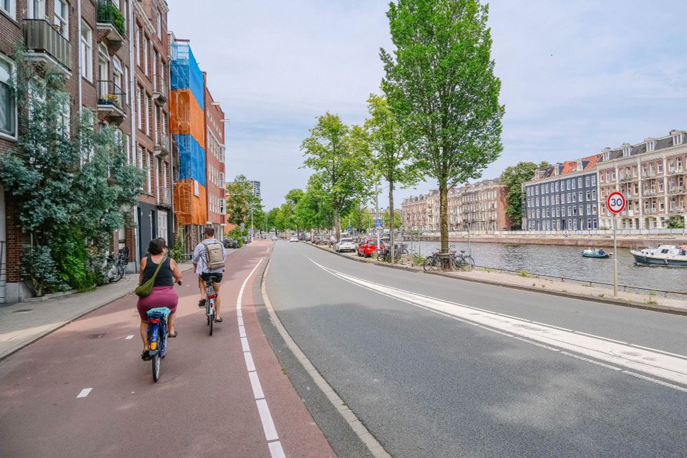 People of all ages and abilities walk, cycle, and drive along the low-traffic streets of Amsterdam, which are lined with buildings, trees, bike infrastructure, and prominent 30 km/hr signage.