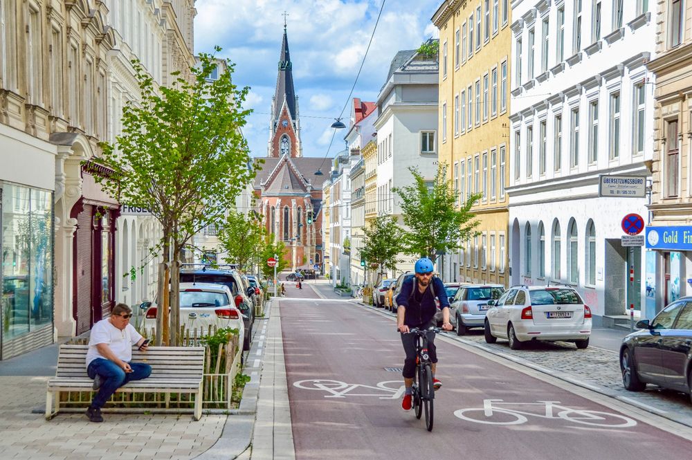 People in casual spring attire cycle on Argentinierstraße in Vienna—a red asphalt “bicycle street” lined with four-storey buildings, trees and plants, and wooden benches. 