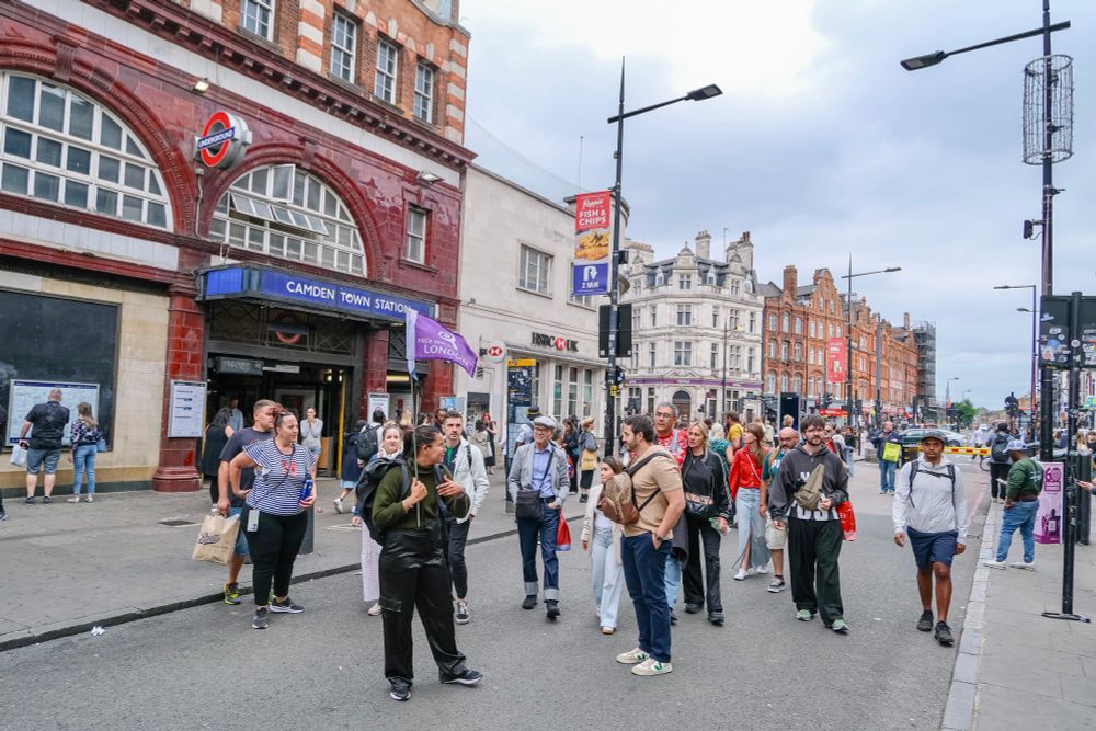 People walk, cycle, sit, and talk along a temporarily car-free stretch of Camden High Street in North London, which is lined with retail shopfronts, trees and flowers, terraces, bike parking, and benches.