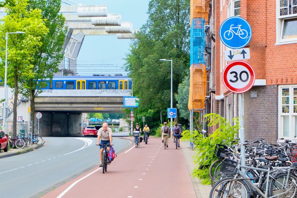 People of all ages and abilities walk, cycle, and drive along the low-traffic streets of Amsterdam, which are lined with buildings, trees, bike infrastructure, and prominent 30 km/hr signage.