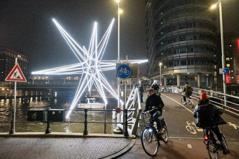 Cyclists travel across a car-free bridge in the harbour of Amsterdam. A neon light art installation sits on top the water.
