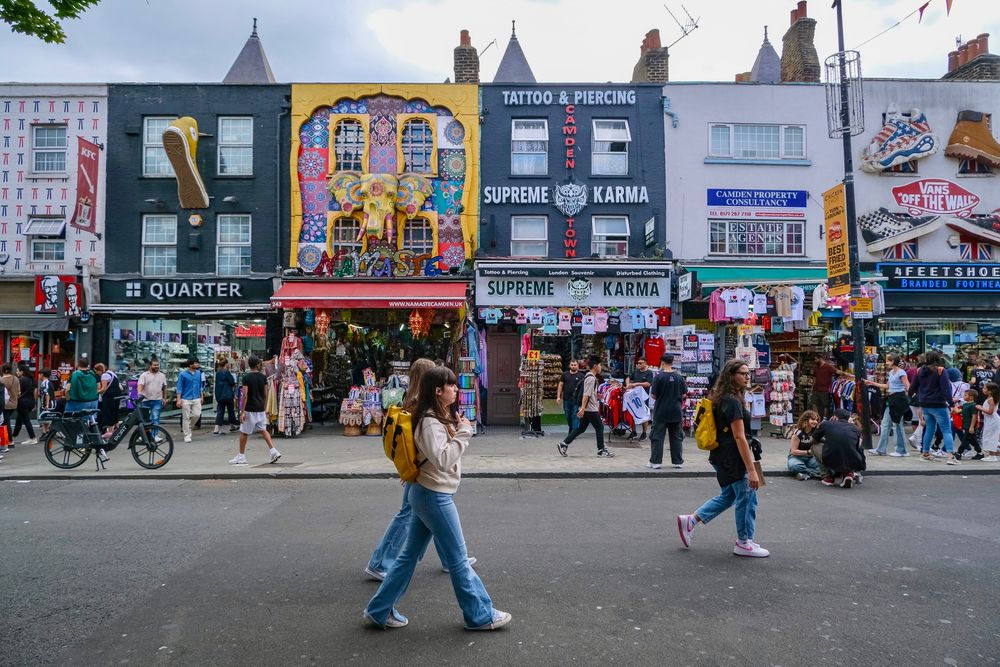 People walk, cycle, sit, and talk along a temporarily car-free stretch of Camden High Street in North London, which is lined with retail shopfronts, trees and flowers, terraces, bike parking, and benches.