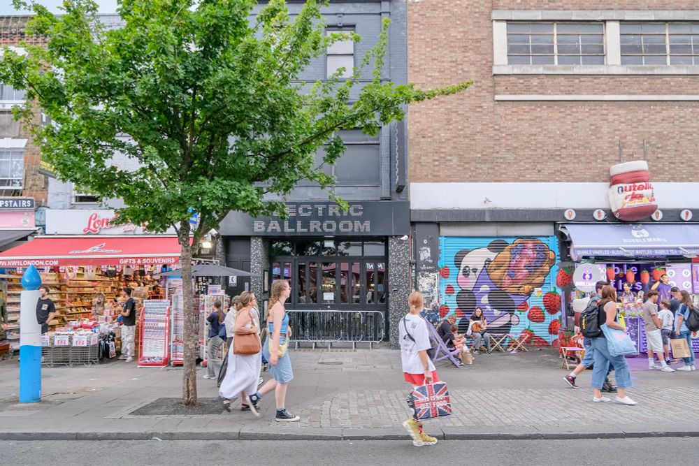 People walk, cycle, sit, and talk along a temporarily car-free stretch of Camden High Street in North London, which is lined with retail shopfronts, trees and flowers, terraces, bike parking, and benches.