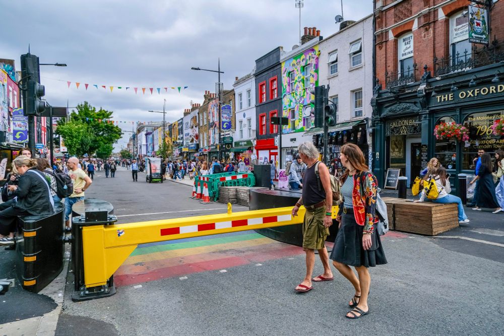 People walk, cycle, sit, and talk along a temporarily car-free stretch of Camden High Street in North London, which is lined with retail shopfronts, trees and flowers, terraces, bike parking, and benches.