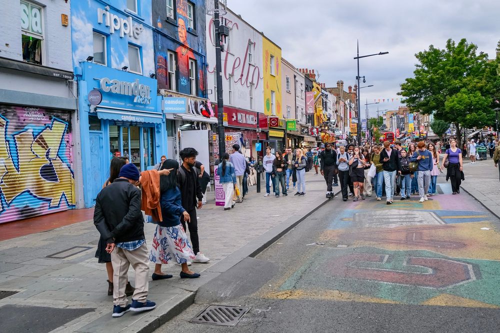 People walk, cycle, sit, and talk along a temporarily car-free stretch of Camden High Street in North London, which is lined with retail shopfronts, trees and flowers, terraces, bike parking, and benches.