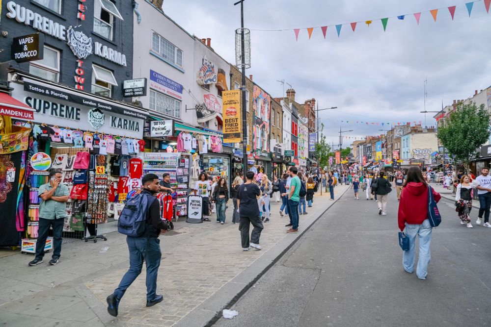 People walk, cycle, sit, and talk along a temporarily car-free stretch of Camden High Street in North London, which is lined with retail shopfronts, trees and flowers, terraces, bike parking, and benches.