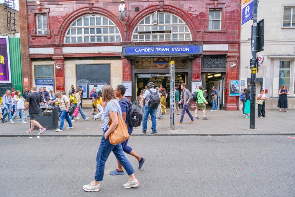 People walk, cycle, sit, and talk along a temporarily car-free stretch of Camden High Street in North London, which is lined with retail shopfronts, trees and flowers, terraces, bike parking, and benches.