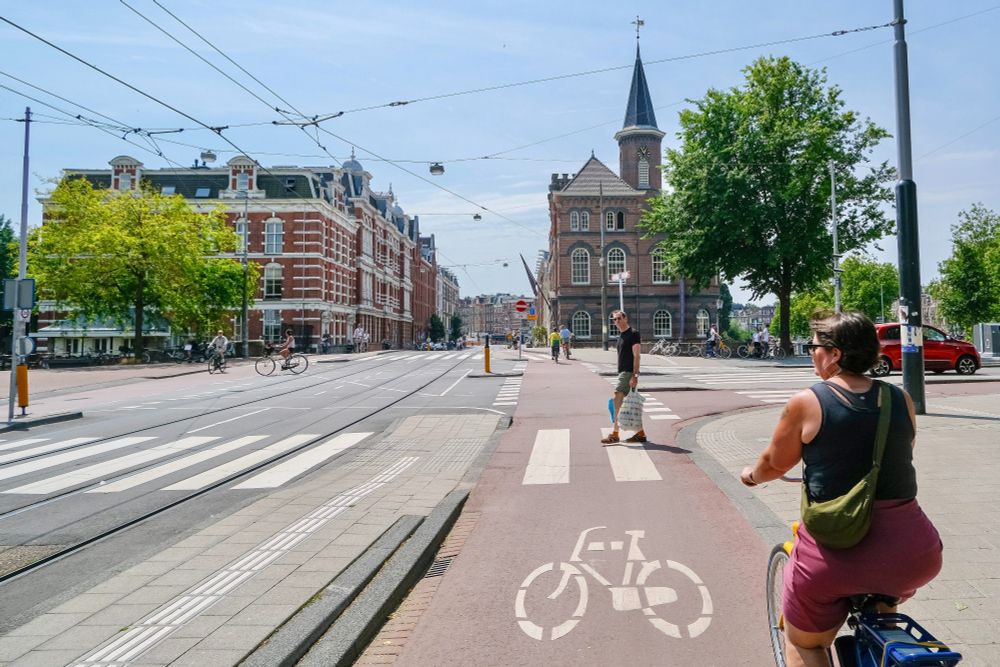 People of all ages and abilities walk, cycle, and drive along the low-traffic streets of Amsterdam, which are lined with buildings, trees, bike infrastructure, and prominent 30 km/hr signage.