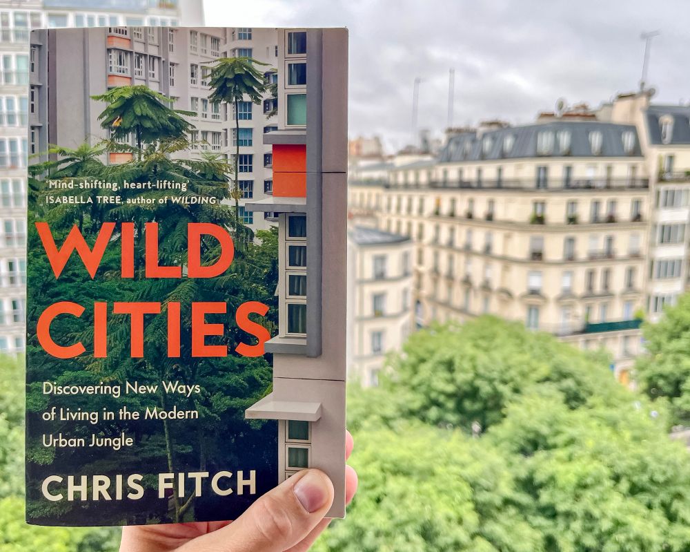 A hand holds a paperback book, “Wild Cities” by Chris Fitch, in front of a Parisian street lined with trees and high rise apartment buildings.