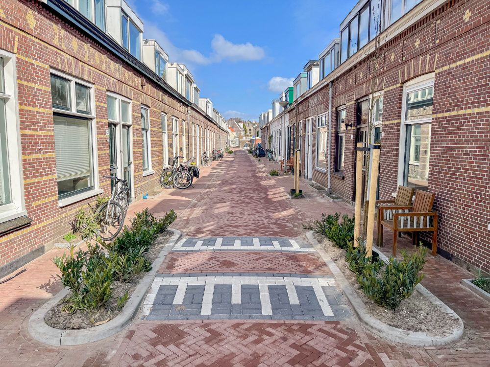People walk along a “woonerf” or “living street” lined with trees, plants, lampposts, benches, and two-storey brick buildings in Delft, The Netherlands.